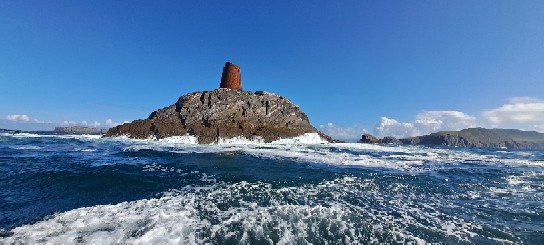 Photo of the remains of a steel lighthouse swept away in a storm C1870, on Calf Rock set in ocean swells with a sunny blue sky.